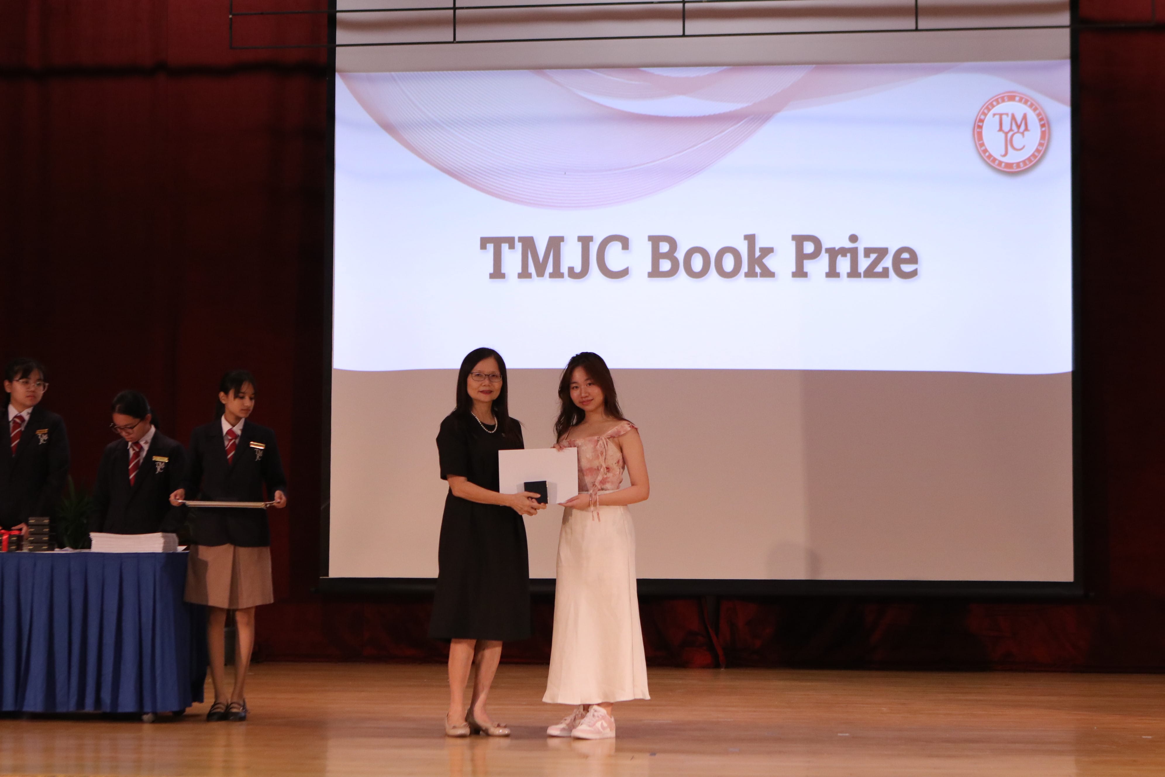 A woman in black presenting the TMJC Book Prize certificate and gift to a student on stage.